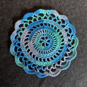 May include: A handmade, circular crochet doily in shades of blue and green. The doily features a scalloped edge and an open, lacy design. The center has a star-like pattern. The color gradient goes from dark blue to light blue and green.
