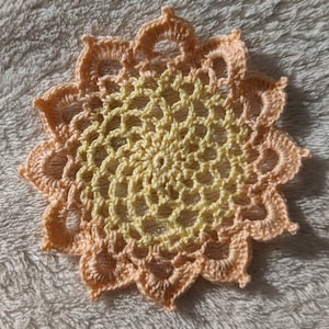 May include: A crocheted doily with a yellow center and peach outer edges. The doily is circular and has a delicate, lacy design.