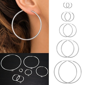 May include: A collection of silver-toned hoop earrings in various sizes, featuring a thin, circular wire design. The set includes multiple pairs of hoops, ranging from small to large, displayed both individually and as worn on an earlobe.