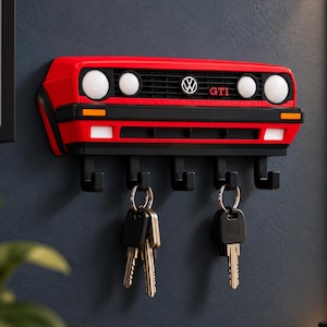 Könnte beinhalten: Ein roter Wand-Schlüsselhalter in Form des Frontgrills eines Volkswagen Golf GTI. Das Design zeigt das ikonische VW-Logo, den GTI-Schriftzug, runde Scheinwerfer und fünf schwarze Haken an der Unterseite, an denen zwei Schlüsselbunde hängen.
