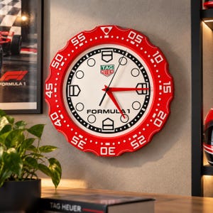 Op de afbeelding: Een ronde wandklok met een rode, geschulpte rand met witte minuutmarkeringen van 05 tot 55. De witte wijzerplaat toont het TAG Heuer-logo, de tekst FORMULA 1, zwarte uurmarkeringen en rode wijzers in een horlogestijl.
