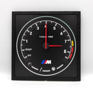 Op de afbeelding: Een zwart-witte klok met een rode en gele wijzer, ontworpen om eruit te zien als een toerenteller van een auto. De wijzerplaat van de klok heeft het BMW-logo en de tekst "1/min x 1000" en "READY OFF".