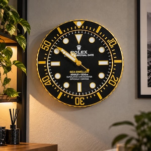 Könnte beinhalten: Eine runde Wanduhr mit schwarzem Zifferblatt und goldfarbener Lünette. Das Zifferblatt zeigt weiße runde und rechteckige Stundenmarkierungen, goldene Zeiger sowie den Text ROLEX OYSTER PERPETUAL DATE SEA-DWELLER 4000ft=1220m SUPERLATIVE CHRONOMETER OFFICIALLY CERTIFIED.