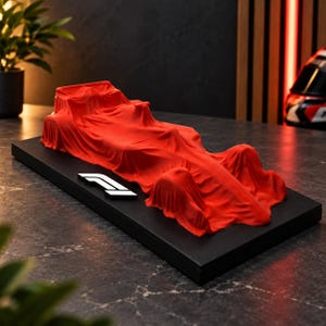 May include: A model of a Formula 1 race car covered by a vibrant red fabric drape, positioned on a rectangular black display base. The base features the white F1 logo on the left side, showcasing the silhouette of the car beneath the cloth.