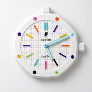 May include: A white octagonal wall clock featuring a textured grid dial with the text AP x Swatch and Royal Pop. The clock face displays colorful hour markers in orange, blue, purple, yellow, black, and green, with light blue hour and minute hands.