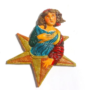 May include: A decorative wall plaque featuring a sculpted figure with brown hair, wearing a blue and red garment, emerging from a large, five-pointed gold glitter star. The figure has a hand placed on their chest and looks upward.