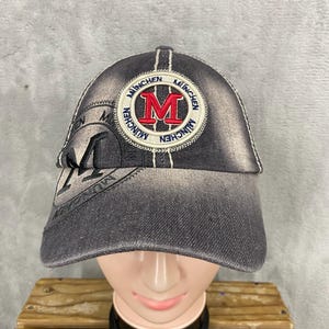 May include: A denim baseball cap with a faded gray wash. The cap features a circular patch with the word "MUNCHEN" in a circle and a large red "M" in the center. White stitching accents the brim and crown.