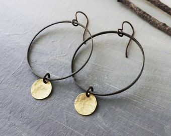 Può includere: Un paio di orecchini a cerchio con ciondoli a disco in oro martellato. I cerchi sono in metallo nero e hanno un design semplice e minimalista.