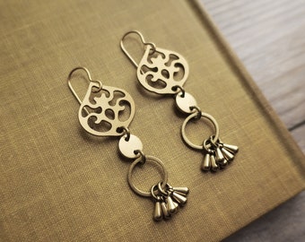 May include: A pair of gold-colored dangle earrings. Each earring features a decorative filigree top, a small disc, a ring, and a cluster of small, teardrop-shaped charms. The earrings are displayed on a textured, tan surface.
