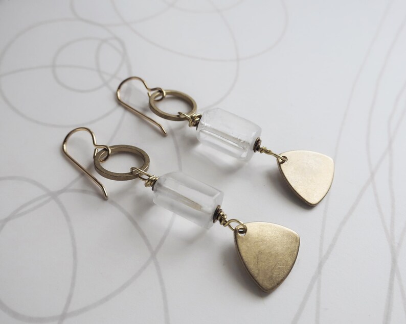 May include: A pair of gold-colored earrings with clear, rectangular beads and small, gold-colored, triangular pendants. The earrings have a simple design with a hook closure and a circular accent.