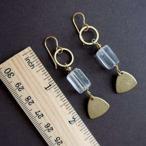 May include: A pair of gold-colored earrings with clear, rectangular beads and small, brass-colored triangle pendants. The earrings are displayed on a dark background next to a wooden ruler.