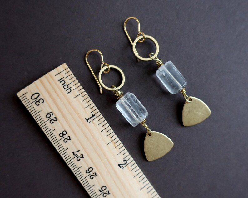 May include: A pair of gold-colored earrings with clear, rectangular beads and small, brass-colored triangle pendants. The earrings are displayed on a dark background next to a wooden ruler.