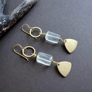 May include: A pair of gold-colored earrings with a light blue, rectangular bead and a gold-colored, triangular charm. The earrings have a simple hook design and are displayed on a dark gray surface.