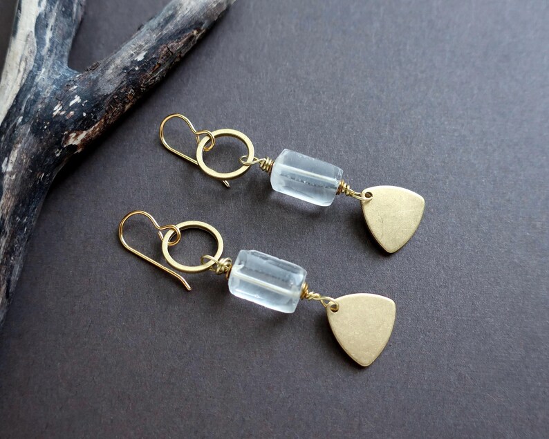 May include: A pair of gold-colored earrings with a light blue, rectangular bead and a gold-colored, triangular charm. The earrings have a simple hook design and are displayed on a dark gray surface.