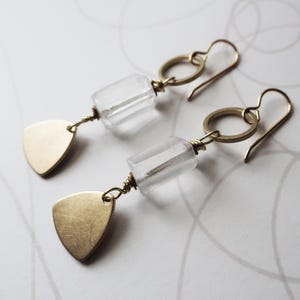 May include: A pair of gold-toned earrings. Each earring features a clear, rectangular bead, a gold ring, and a gold triangle pendant. The earrings have a simple, elegant design, suitable for everyday wear or special occasions.
