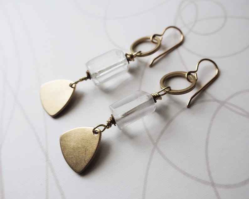 May include: A pair of gold-toned earrings. Each earring features a clear, rectangular bead, a gold ring, and a gold triangle pendant. The earrings have a simple, elegant design, suitable for everyday wear or special occasions.