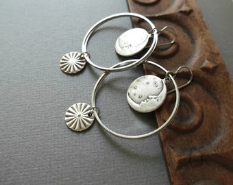 May include: A pair of silver hoop earrings. Each earring has a circular pendant with a sunburst design and a larger, round pendant with a textured moon and star design. The earrings are displayed on a dark gray surface.