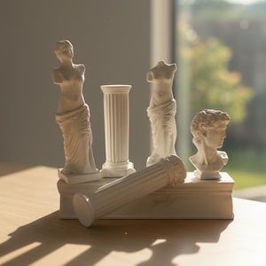 Puede incluir: Una colección de pequeñas esculturas clásicas blancas, que incluyen bustos, figuras parciales y columnas. Las esculturas están dispuestas sobre una base rectangular, con una columna tumbada de lado. La escena está iluminada por luz natural.