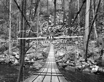 May include: A black and white photograph of a wooden bridge over a stream in a woodland. The bridge is constructed from wooden planks and is supported by cables. The bridge is surrounded by trees and rocks.