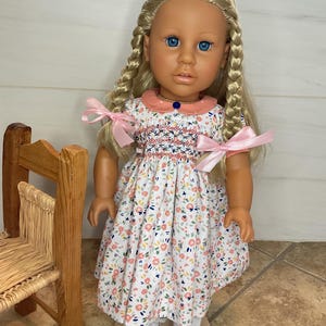 May include: A doll with blonde hair styled in two braids, blue eyes, and a light skin tone. The doll wears a white dress featuring a multi-colored floral pattern, a pink collar with white dots, and pink ribbons tied at the sleeves, paired with white sneakers.