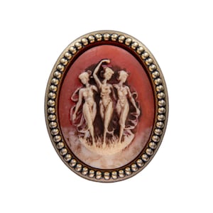 May include: An oval-shaped cameo brooch with a gold-toned metal frame and a red cameo featuring three women in white relief.