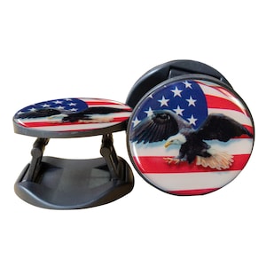 May include: A black phone grip with a foldable stand features a printed image of an American bald eagle flying over an American flag with red and white stripes and blue stars.