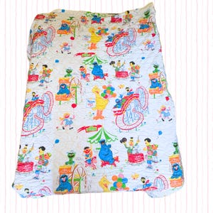 May include: White quilted blanket featuring a colorful Sesame Street theme park design. The blanket showcases roller coasters, characters like Big Bird, Cookie Monster, and Elmo, and various carnival elements. The design is repeated across the blanket.