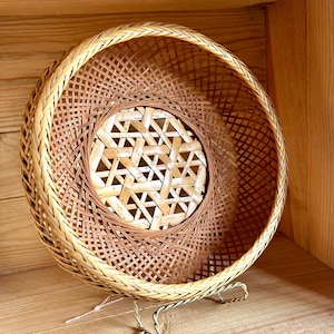May include: A round, woven bamboo basket with a spiral design. The basket features a light brown and tan color scheme, with a geometric pattern at the center. The basket is displayed on a metal stand.