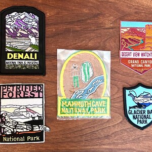 May include: A collection of embroidered patches featuring national park designs. Patches include Denali, Petrified Forest, Desert View Watchtower, Mammoth Cave, and Glacier Bay National Park. Each patch showcases unique landscape illustrations and park names.