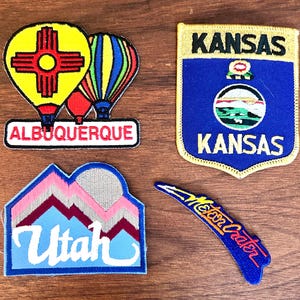 May include: Four embroidered patches featuring travel destinations. One patch displays two hot air balloons with "Albuquerque" below. Another is a blue shield with "Kansas" and a landscape. A third patch shows mountains and "Utah". The last is a blue patch with "Meteor Crater".