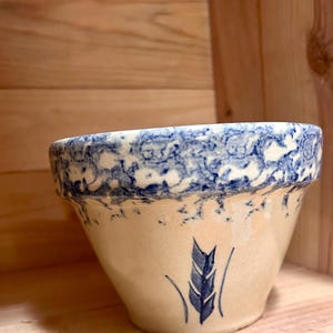 May include: A cream-colored ceramic bowl with a blue and white speckled rim. The bowl features a blue wheat stalk design on the front. The bowl is set against a wooden background.