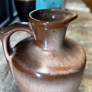 May include: A small brown ceramic pitcher with a white glaze. The pitcher has a rounded body and a small handle.