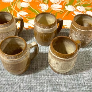May include: Five brown ceramic mugs with a barrel-like design. Each mug has a handle and a light-colored band around the middle. The mugs are arranged on a gray and white patterned surface, with an orange floral background.