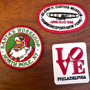 May include: Three embroidered patches on a wooden surface. One patch features Santa Claus with text "Santa's Workshop, North Pole, NY." Another is an oval patch with text "Glenn H. Curtiss Museum, June Bug 1903, Hammondsport, New York." The third patch says "Love Philadelphia."