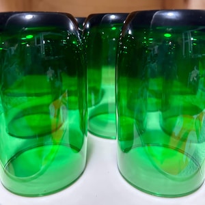 May include: A close-up shot of several green-tinted glass tumblers. The glasses have a rounded shape with a darker green rim. The clear glass allows light to pass through, creating reflections and highlights.