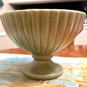 May include: A light green, pedestal bowl with a ribbed exterior. The bowl has a wide, rounded shape and sits on a short, circular base. The bowl is made of ceramic or stoneware and has a speckled texture.