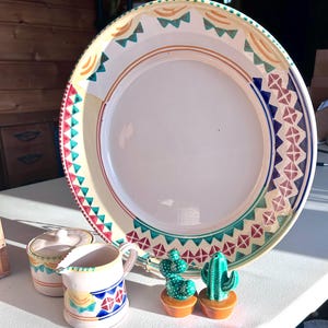 May include: A large, decorative ceramic plate with a cream-colored center and a colorful border featuring geometric patterns in red, teal, and blue. Two small cactus figurines in orange pots and a small creamer and sugar bowl are also present.