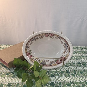 May include: A white ceramic bowl with a scalloped edge and a decorative rim featuring a detailed pattern of red roses, blue flowers, and brown scrollwork. The bowl is displayed on a gold metal stand, showcasing its intricate floral border design.