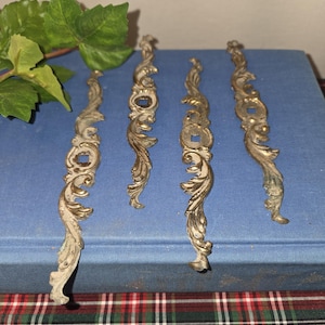 May include: Four antique brass drawer pulls with ornate, scrolling designs. The pulls have a weathered, aged appearance with a mix of gold and silver tones. Each pull is approximately 6 inches long, featuring decorative details.