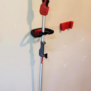 Op de afbeelding: Een rood-zwarte Milwaukee FUEL-grastrimmer aan de muur, met een bijpassende rode muurbevestiging. De trimmer heeft een zilveren schacht en een zwarte handgreep.