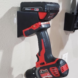 May include: Black and red cordless drill with a black battery pack mounted on a wall. The drill is a Milwaukee M18 model. The drill is mounted on a black holder.