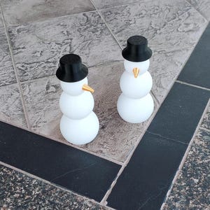 Könnte beinhalten: Zwei weiße Schneemänner mit schwarzen Zylindern und goldenen Nasen. Die Schneemänner stehen auf einem gefliesten Boden.