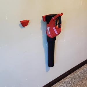 May include: A red and black Milwaukee leaf blower mounted on a wall, with a separate red mounting bracket. The blower has a black nozzle and a red handle. The wall is light beige.