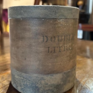 May include: A cylindrical container with a distressed look. The container is mainly brown with darker bands at the top and bottom. The text "DOUBLE LITRES" is printed on the side. The container appears to be made of wood or a similar material.