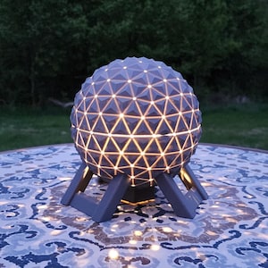 Può includere: Una lampada sferica geometrica grigia con una luce bianca che brilla attraverso i fori. La lampada è su un tavolo rotondo con una superficie a motivi.