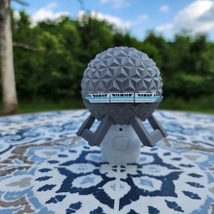 Op de afbeelding: Een grijs, geometrisch bolvormig object met een miniatuur monorail ontwerp. De bol staat op een witte basis met grijze steunen. De achtergrond toont een vage groene landschap en een blauwe lucht, wat wijst op een buitenomgeving.