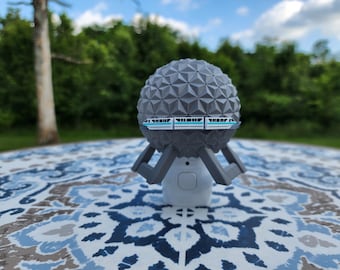Könnte beinhalten: Ein graues, geometrisches kugelförmiges Objekt mit einem Miniatur-Monorail-Design. Die Kugel sitzt auf einem weißen Sockel mit grauen Stützen. Der Hintergrund zeigt eine verschwommene grüne Landschaft und einen blauen Himmel, was auf eine Umgebung im Freien hindeutet.