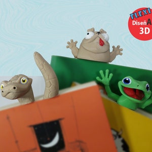 May include: Three 3D printed bookmark figurines: a green frog with its mouth open, a brown snake with its head peeking out from behind a book, and a brown pug with its tongue sticking out. The bookmarks are on top of a stack of books.