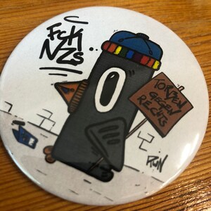 Könnte beinhalten: Ein schwarz-weißer Cartoon-Pinguin, der einen blauen und regenbogenfarbenen Hut trägt und ein Schild mit der Aufschrift "Torben gegen Rechts" hält. Der Pinguin geht auf einem Bürgersteig mit einer Backsteinmauer im Hintergrund. Der Text "Fck NZS" steht in der oberen linken Ecke des Bildes.