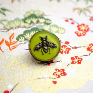Puede incluir: Un botón redondo con un centro verde lima y un emblema de abeja en relieve de color bronce metálico. El botón tiene un borde de metal color bronce oscuro y un acabado liso y brillante, mostrando detalles en las alas y el cuerpo del insecto.